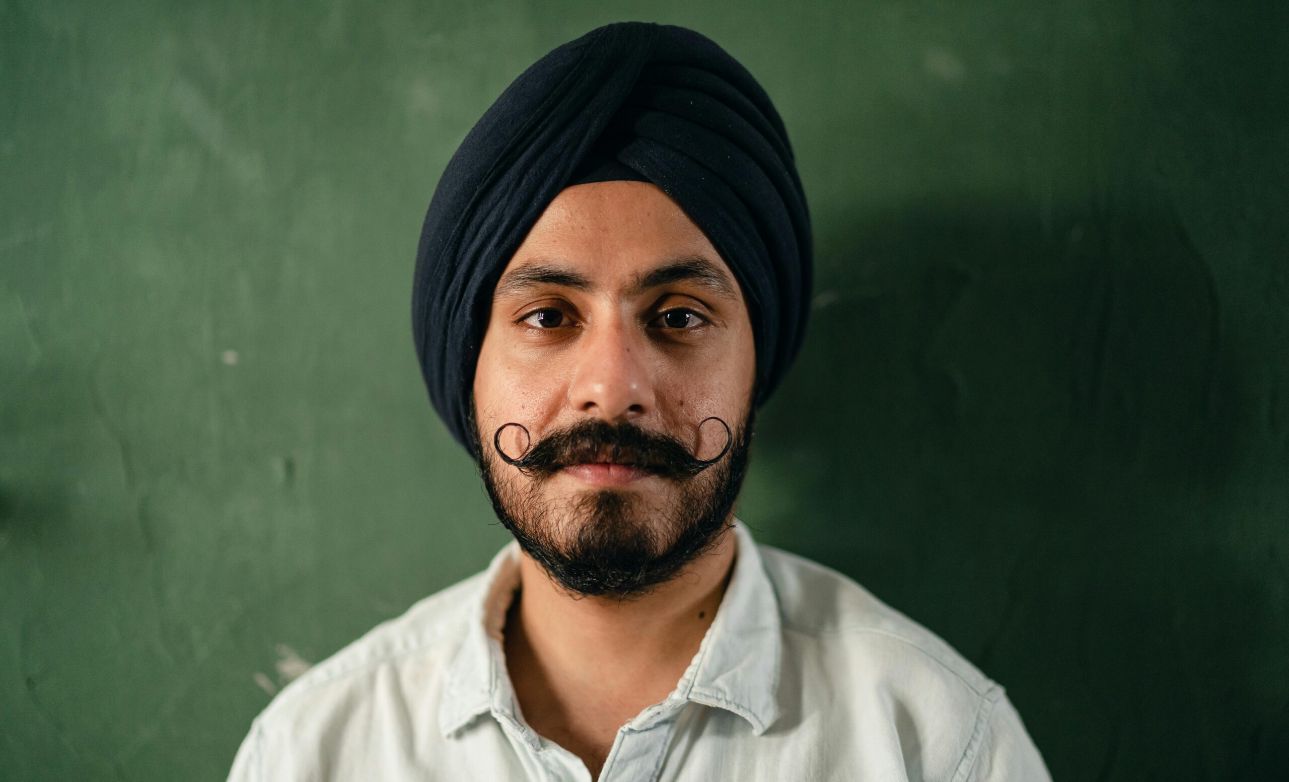 Close-up portrait of a young South Asian man with a turban and mustache against a green background.